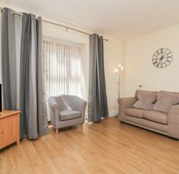 A living room with a television on a wooden stand a grey armchair grey curtains a two-seater sofa three cushions and a wall clock at 21 Eamont Park Penrith