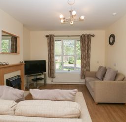A living room with two beige sofas a wall clock a TV on a glass stand a fireplace and a window with curtains at 12 Eamont Park in Penrith