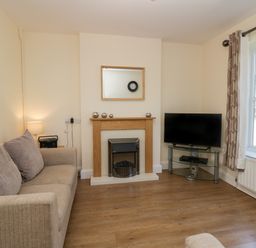A living room with a beige sofa a fireplace a television on a glass stand and a window with curtains at 12 Eamont Park in Penrith