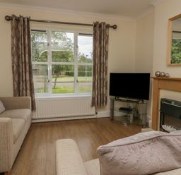 A living room with two sofas a television on a glass stand a fireplace and a large window at 9 Eamont Park in Penrith