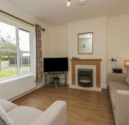 A living room with two beige sofas a television on a glass stand a fireplace and a window with curtains at 9 Eamont Park in Penrith