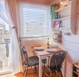 A small dining table with two chairs and a bottle of wine near a window with blinds and shelves at Sea Spray in Lyme Regis