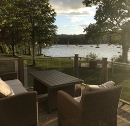 An outdoor seating area with a table and cushioned chairs overlooking a lake with boats and trees at Loubi's Lakeside Lodge M1 Fallbarrow Park near Bowness-On-Windermere