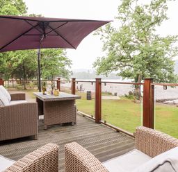 An outdoor deck with a wicker sofa and chairs a table with glasses and bottles under a large umbrella overlooking a lakeside park at Loubi's Lakeside Lodge M1 Fallbarrow Park near Bowness-On-Windermere