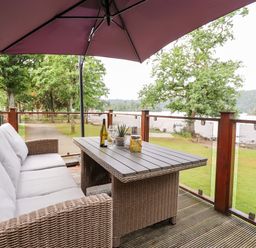 An outdoor seating area with a wicker sofa and table under a large umbrella next to a lake at Loubi's Lakeside Lodge M1 Fallbarrow Park near Bowness-On-Windermere