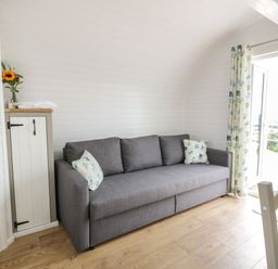 A small living area with a gray sofa and floral cushions next to a wooden cabinet and glass door with floral curtains at Laird Lodge 3 in Lochmaben