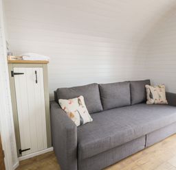A gray sofa with patterned pillows next to a small cabinet with towels on top in a room with white wooden walls and wood flooring at Laird Lodge 4 in Lochmaben