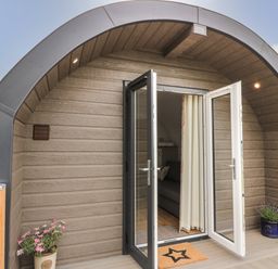 The entrance of a curved wooden structure with open glass doors and flower pots on each side at Laird Lodge 4 in Lochmaben