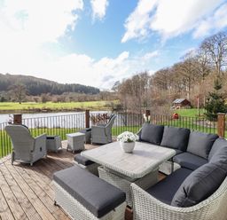 An outdoor deck with wicker furniture including chairs and a table overlooking a lake and trees at Tan Yr Allt in Dolanog near Meifod Powys