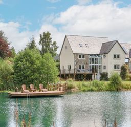 A house with a wooden deck and outdoor chairs by a pond surrounded by trees at Tan Yr Allt in Dolanog near Meifod Powys
