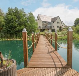 A wooden bridge over a pond leading to a house surrounded by trees and plants at Tan Yr Allt in Dolanog near Meifod Powys