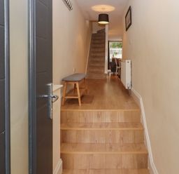 A hallway with wooden stairs leading to a carpeted staircase a bench on the left wall a radiator on the right wall and a dining area visible at the end at Mimosa in Dawlish