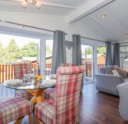 A dining area with a glass table and plaid chairs next to a living room with gray sofas and curtains at Fell Foot Lodge in Fellside 9 Limefitt Park near Troutbeck