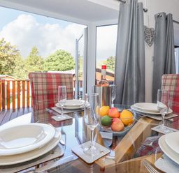 A dining table set with white plates glasses a bowl of fruit and a bottle of wine next to plaid chairs and glass doors at Fell Foot Lodge in Fellside 9 Limefitt Park near Troutbeck