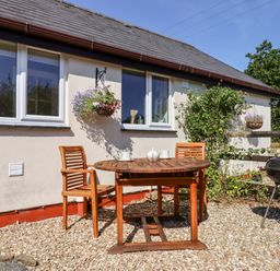 A patio area with a round wooden table and two wooden chairs next to a house with two windows and hanging flower baskets at Little Rowan at Rowan Park Farm in Ashwater