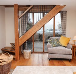 A living room with a gray armchair with pillows near wooden stairs with a basket of logs and a side table lamp at Little Orchard in Ashburton Down near Ashburton