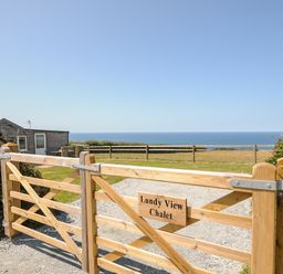 A wooden gate with a sign reading Lundy View Chalet overlooking a field and ocean in Widemouth Bay