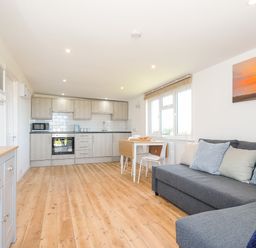 A kitchen and living room area with wooden floors a grey sofa with pillows a dining table and chairs and kitchen cabinets at Lundy View Chalet in Widemouth Bay