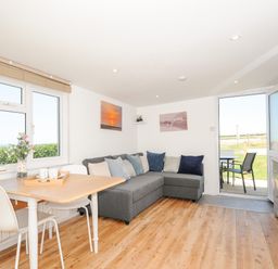 A living room with a dining table and chairs a gray corner sofa and an open door leading outside at Lundy View Chalet in Widemouth Bay