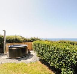 An outdoor hot tub on gravel near a wooden fence with a hedge and sea view at Lundy View Chalet in Widemouth Bay