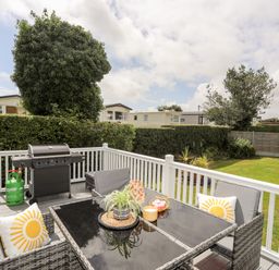 An outdoor patio with a glass table and chairs with cushions and a barbecue grill in a grassy backyard at Moondunes - Pwllheli in Chwilog