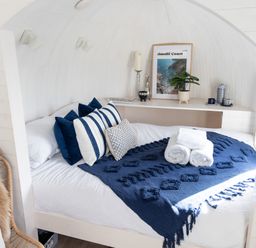 A bed with blue and white pillows and a blue blanket with rolled white towels near a shelf with a plant and picture at Andrus - Mayflower Meadow in Brandesburton