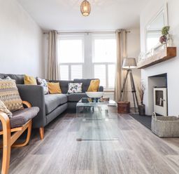 A living room with a gray sectional sofa with cushions a glass coffee table a wooden chair with cushions a floor lamp a fireplace and a basket at Chy An Mor in Mevagissey
