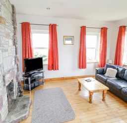 A living room with a stone fireplace a black leather sofa a wooden coffee table a television on a stand and orange curtains at 13 Trawmore Cottages in Keel County Mayo