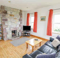 A living room with a stone fireplace a wooden coffee table a black leather sofa and two windows with red curtains at 13 Trawmore Cottages in Keel County Mayo