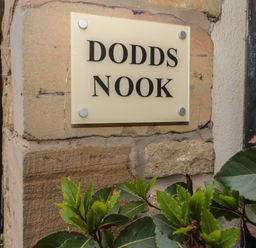A stone wall with a sign reading Dodds Nook and green plant leaves in the foreground at Dodds Nook in Alnwick