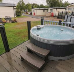A hot tub on a wooden deck with steps surrounded by glass and railing at Waterside Lodge South Lakeland Leisure Village near Carnforth