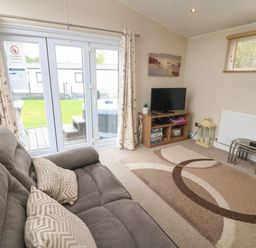 A living room with a grey sofa and patterned cushions a TV on a wooden stand a patterned rug and glass patio doors at Waterside Lodge South Lakeland Leisure Village near Carnforth