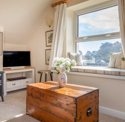 A living room with a wooden chest table with flowers on it a window seat with cushions a television fireplace and framed pictures at Goody in Hope Cove