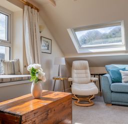 A living area with a wooden chest table with a vase of flowers a white armchair a blue sofa with cushions and a window seat with striped cushions at Goody in Hope Cove