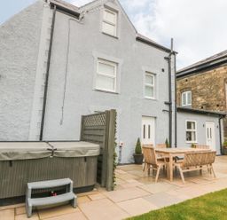 An outdoor patio with a hot tub and wooden dining table with chairs outside a gray house at Fishermans Cottage Malltraeth