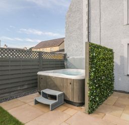 An outdoor hot tub with steps on a patio next to a house with a gray fence and a green privacy screen at Fishermans Cottage in Malltraeth