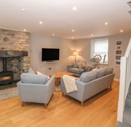 A living room with grey sofas a wooden coffee table a stone fireplace a wall-mounted TV and a staircase at Fishermans Cottage in Malltraeth