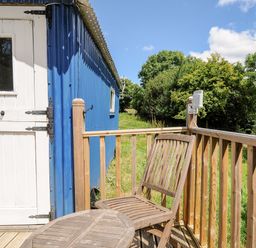 A wooden chair and table on a wooden deck outside a blue building with a white door at Shepherds Hut in Boconnoc near Lostwithiel