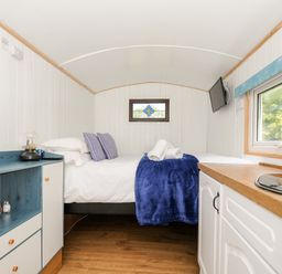 A small room with a bed, pillows, a blue blanket, a cabinet with jars, a sink, and a window at Shepherds Hut Boconnoc near Lostwithiel