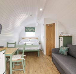 A small room with a bed, a gray sofa, a wooden table with chairs, a wall-mounted TV, and a wooden door at Laird Lodge 5 in Lochmaben