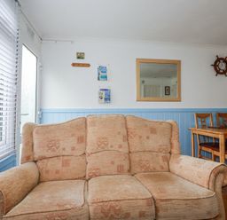 A living room with a beige sofa wooden dining table and chairs a wall mirror and a ship wheel clock at 8 The Park Kilkhampton