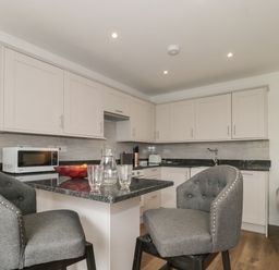A kitchen with white cabinets a microwave on the counter a black countertop with chairs and glassware at The Hideaway in Lympsham near Brean