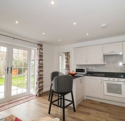 A kitchen with white cabinets black countertop a stove microwave two chairs and glass doors leading to a garden at The Hideaway in Lympsham near Brean