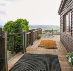 An outdoor wooden deck with a fence a doormat potted flowers and metal table and chairs at Dragonfly Lodge in Worthen near Minsterley