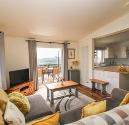 A living room with gray sofas yellow cushions and a wooden coffee table next to an open door to a balcony with a dining table at Dragonfly Lodge Worthen near Minsterley