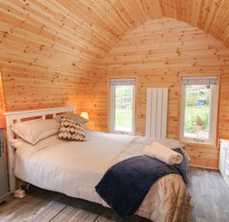 A bedroom with a double bed bedside tables lamps and dresser in a wooden paneled room with two windows at The Pod Worthen near Minsterley