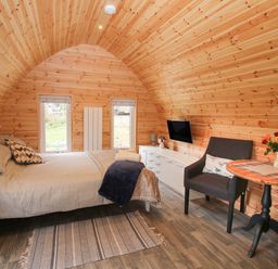 A bedroom with wooden walls a bed two windows a dresser a mounted television two chairs and a small round table at The Pod in Worthen near Minsterley