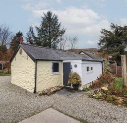 A small stone and wood cottage with a grey door and windows surrounded by gravel and trees at Tyn Y Cefn Near Blaenau Ffestiniog