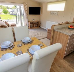 A dining area with a wooden table set for four with white chairs next to a living room with sofas a wall-mounted TV and an open door leading to a garden at Number 15 Carnforth