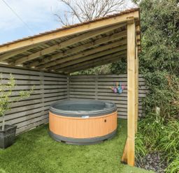 An outdoor covered area with a hot tub artificial grass flooring a potted plant and gardening tools on a gray wooden fence at Orchard Lodge in Llanengan near Abersoch